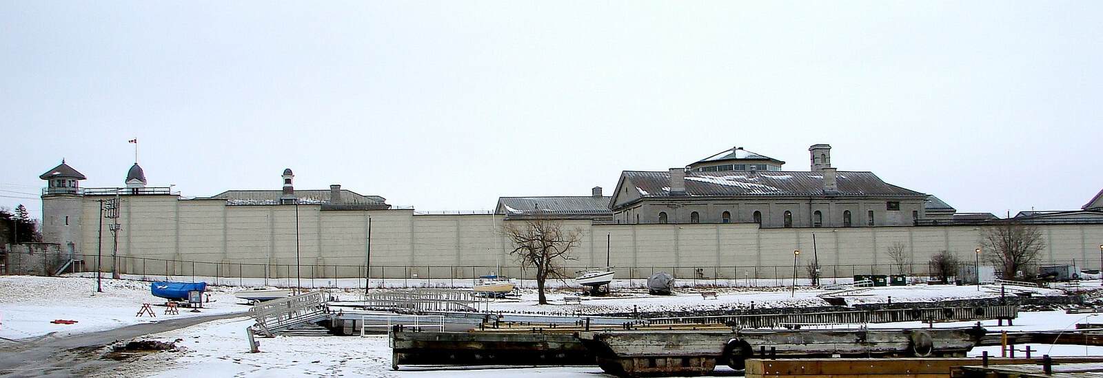 Long limestone perimeter wall and buildings of Kingston Penitentiary beside a snowy waterfront.