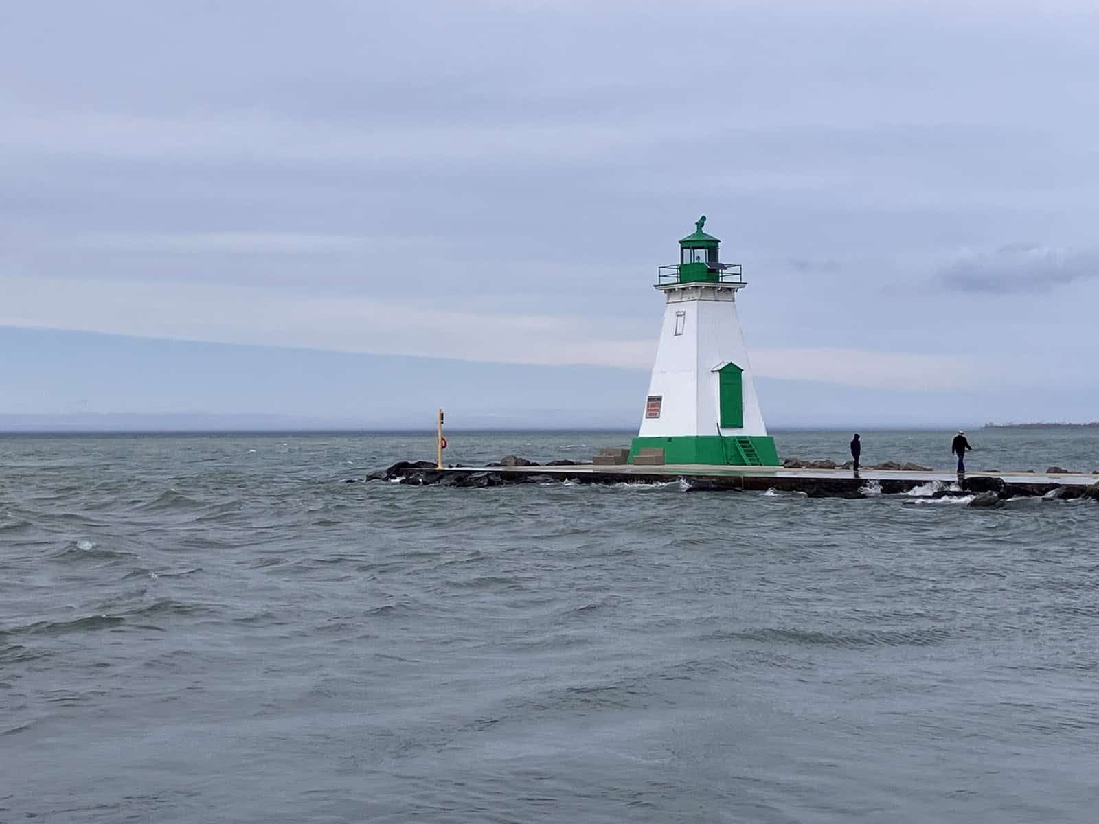 White lighthouse with a green top at the end of a pier on Lake Ontario under a grey sky.