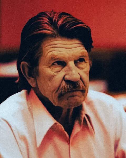 Older man with a moustache in a light shirt seated in a courtroom.