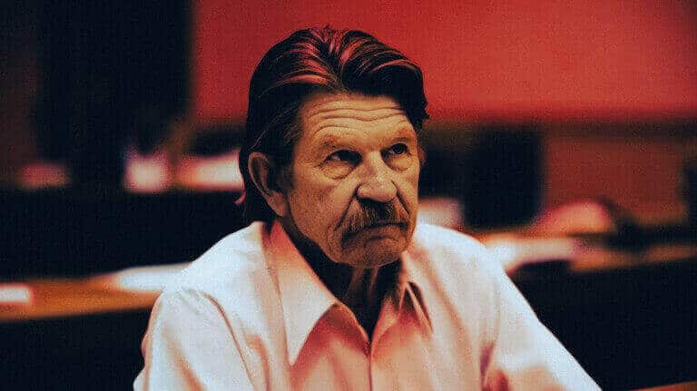 Older man with a moustache in a light shirt seated in a courtroom.