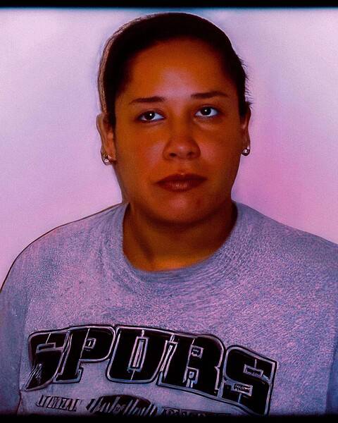 Booking-style photograph of a woman in a gray shirt in front of a pink wall.