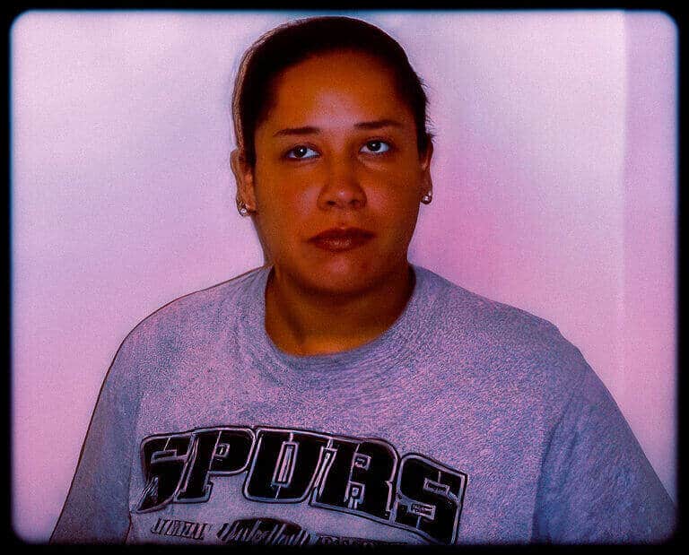 Booking-style photograph of a woman in a gray shirt in front of a pink wall.