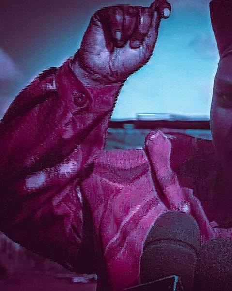 Woman in a headwrap speaking into a microphone with one hand raised, in tinted pink and blue lighting.