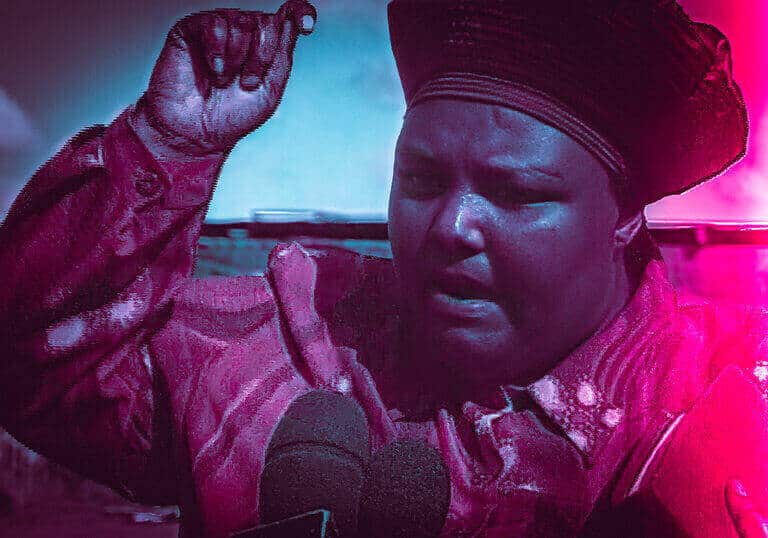 Woman in a headwrap speaking into a microphone with one hand raised, in tinted pink and blue lighting.