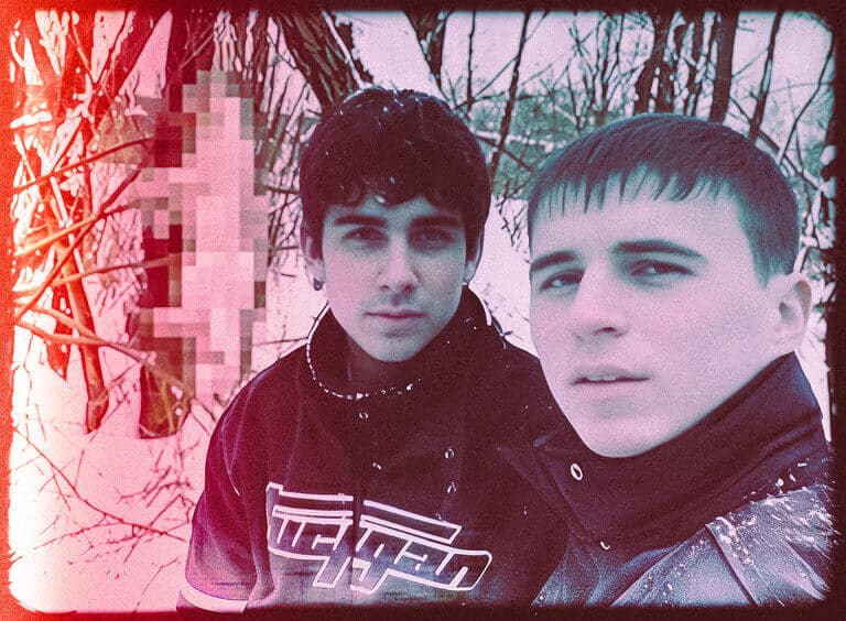 Two young men stand together in a bare winter woodland, with part of the frame pixelated.