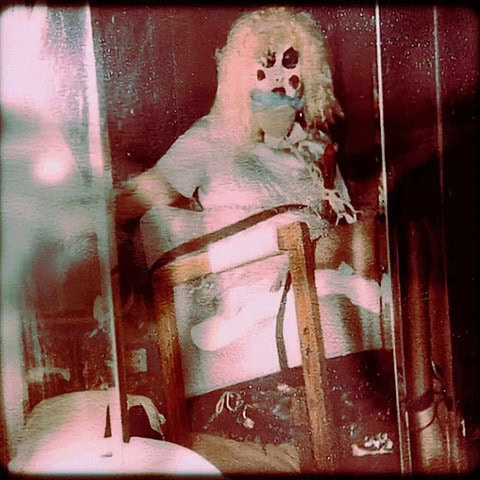 A pale clown figure seated in a red-lit room in a grainy vintage photograph.