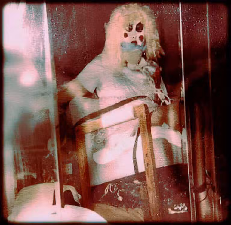 A pale clown figure seated in a red-lit room in a grainy vintage photograph.