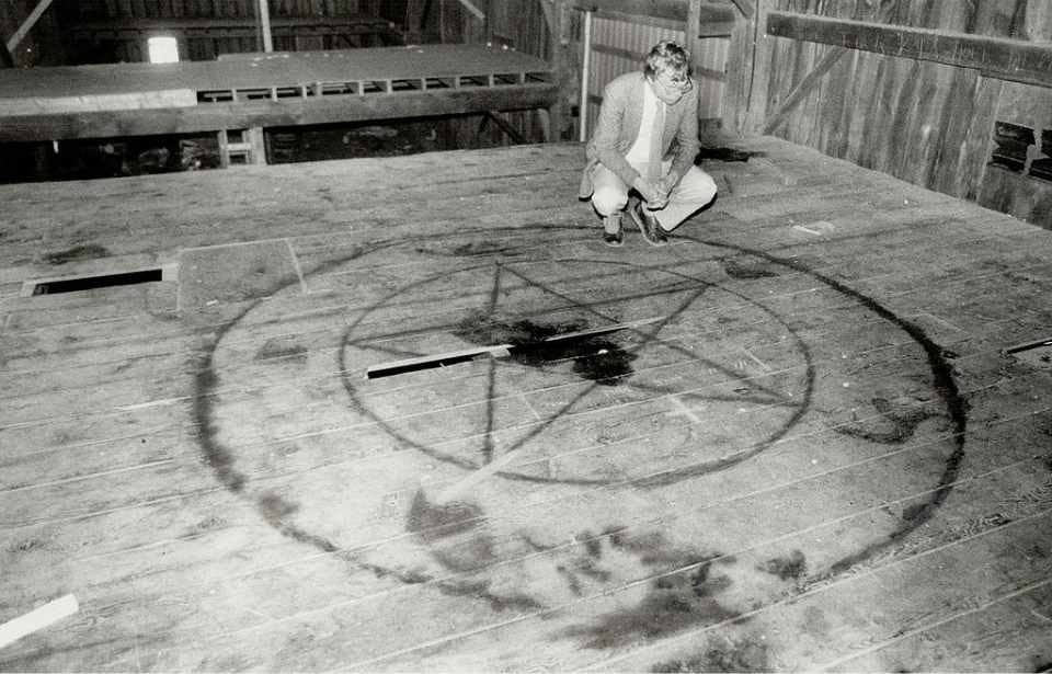 Black-and-white photo of a man crouching beside a large pentagram drawn on the wooden floor of a barn.