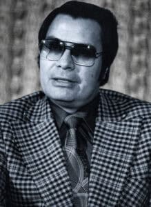 A black and white photograph of Jim Jones wearing dark sunglasses with swept-back dark hair.