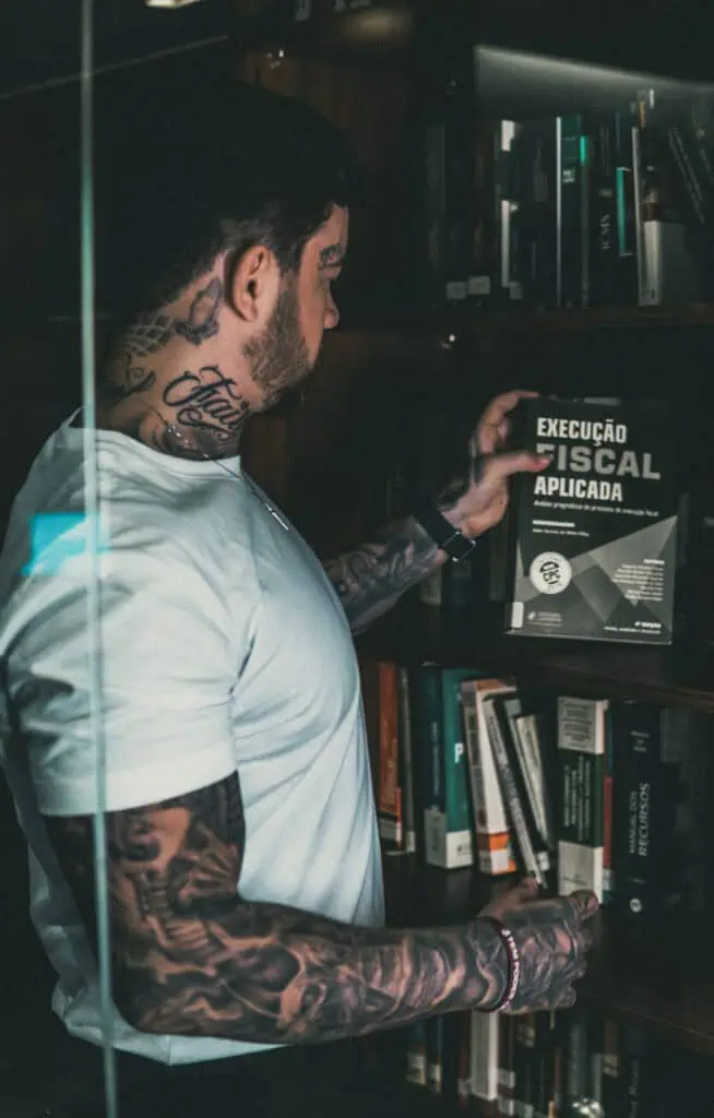 A tattooed man reaches for a Portuguese law textbook on a dark bookshelf.