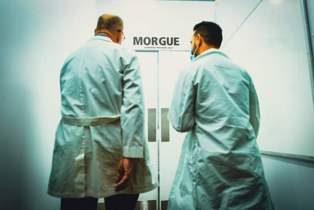 Two medical workers in scrubs stand in front of a hospital morgue door.