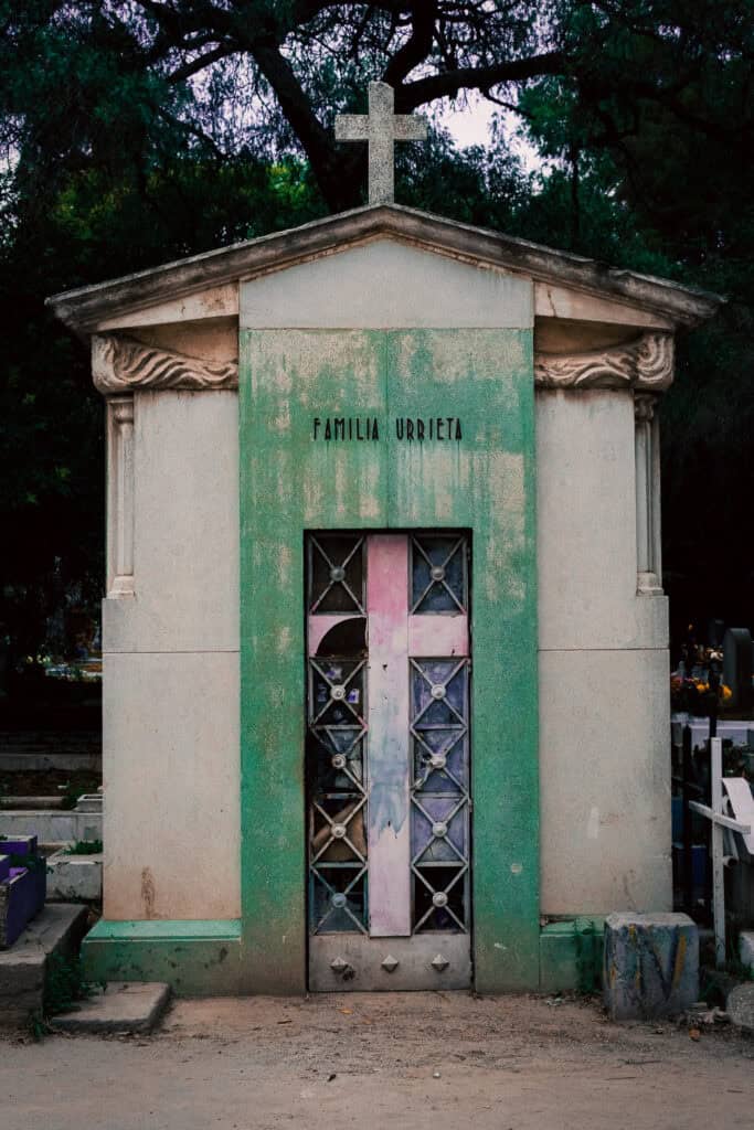 A weathered stone family tomb topped with a cross beneath trees in a cemetery.