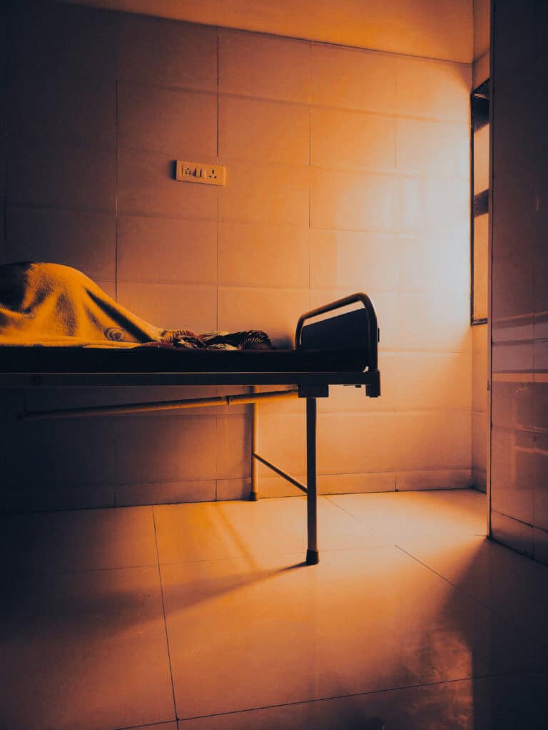 A person resting under a blanket on a metal hospital bed in a dimly lit room.