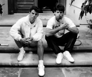 Two young men sitting on steps in casual sportswear, one wearing a Nike jacket, both looking at the camera in a relaxed pose.