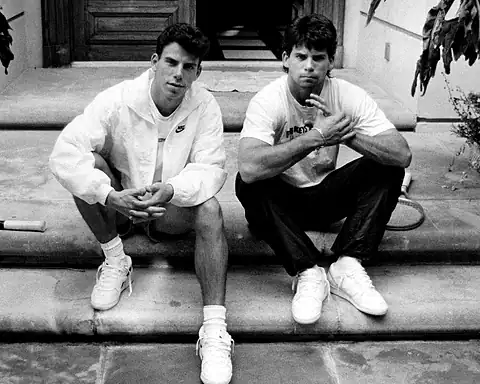 Two young men sitting on steps in casual sportswear, one wearing a Nike jacket, both looking at the camera in a relaxed pose.