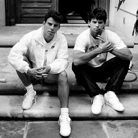 Two young men sitting on steps in casual sportswear, one wearing a Nike jacket, both looking at the camera in a relaxed pose.