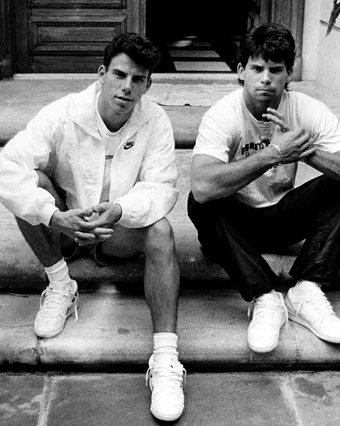 Two young men sitting on steps in casual sportswear, one wearing a Nike jacket, both looking at the camera in a relaxed pose.