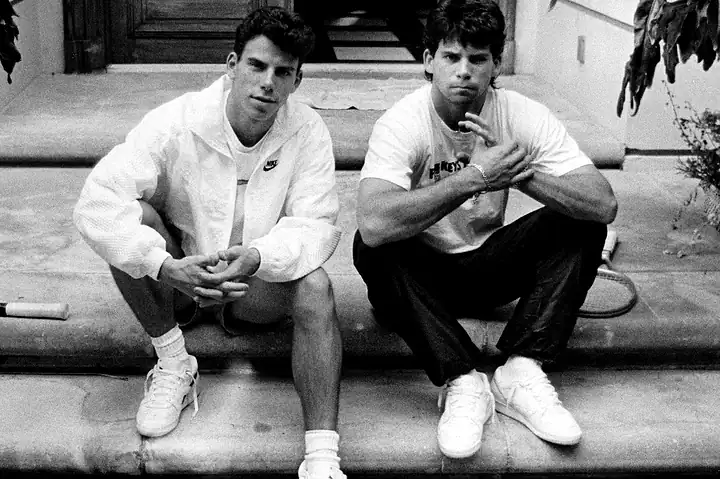 Two young men sitting on steps in casual sportswear, one wearing a Nike jacket, both looking at the camera in a relaxed pose.