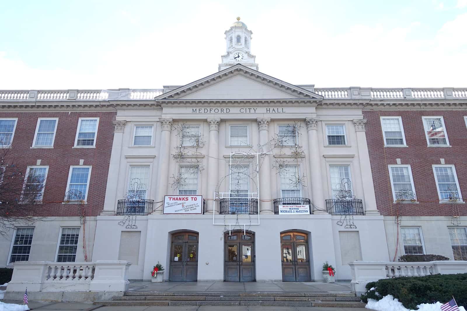 Brick and limestone civic building with a columned entrance and a white cupola under a pale sky.