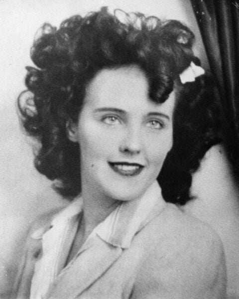 Black and white portrait of Elizabeth Short, a smiling young woman with dark curled hair and a white ornament in her hair.