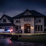 Police cars and yellow tape outside a large suburban house at night.