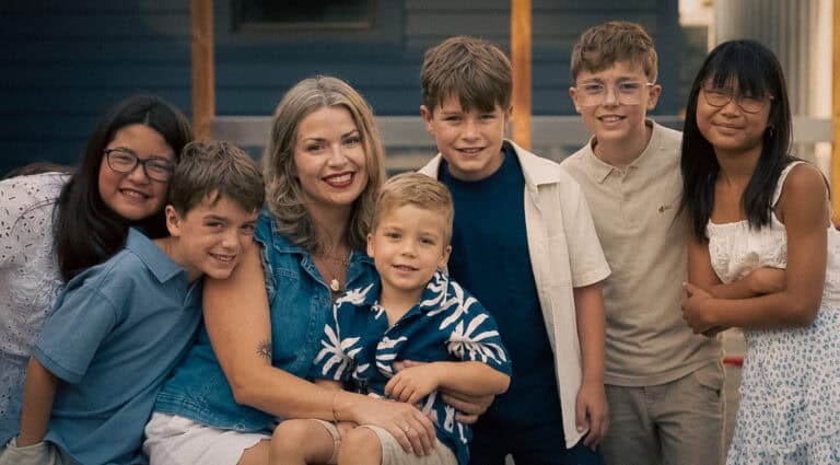A smiling woman surrounded by six children posing together for a family portrait outdoors.