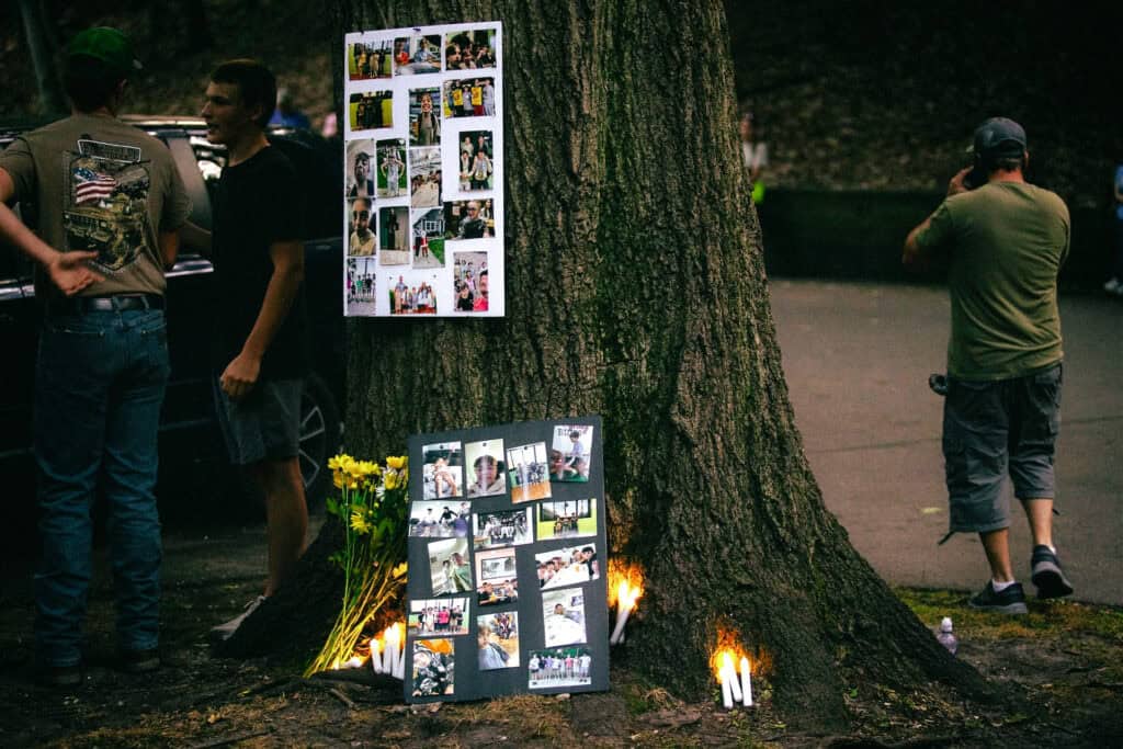 Mourners stand beside photo collage boards and flowers placed against a large tree.