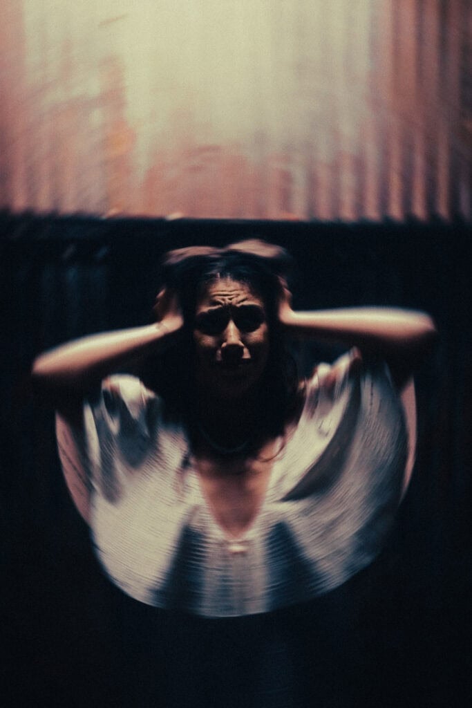 A blurred figure sits in a dim room holding both hands against the sides of their head.