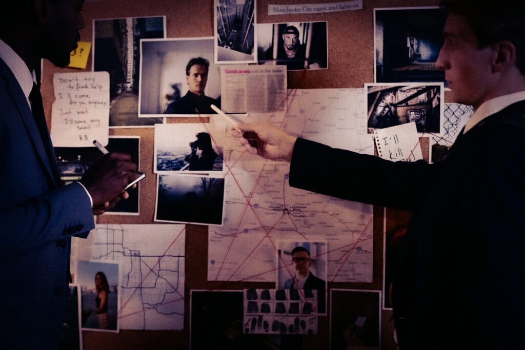 Two detectives in suits study a cork board covered with photographs, notes and a map connected by red string.