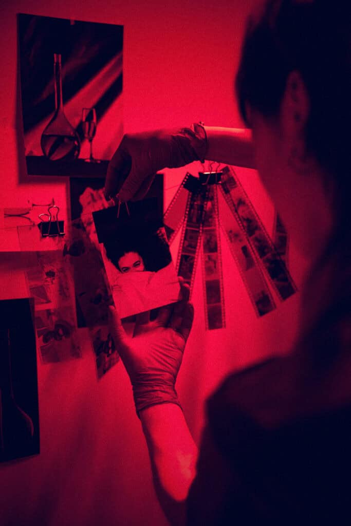 A hand holds a photographic print above film strips and clips on a table in a darkroom lit by red safelight.