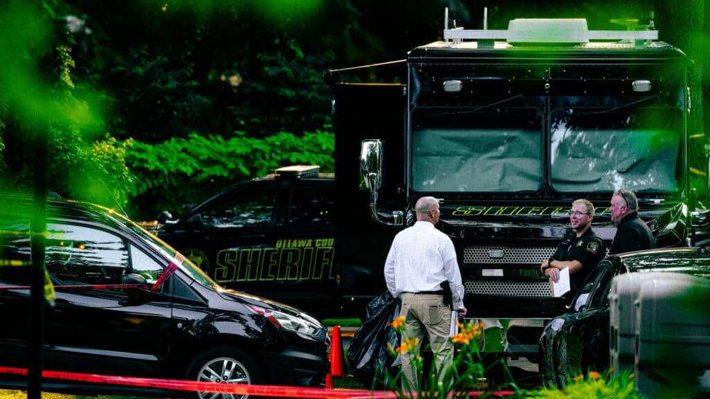 Officers and an investigator stand beside an Ottawa County sheriff command truck behind police tape.
