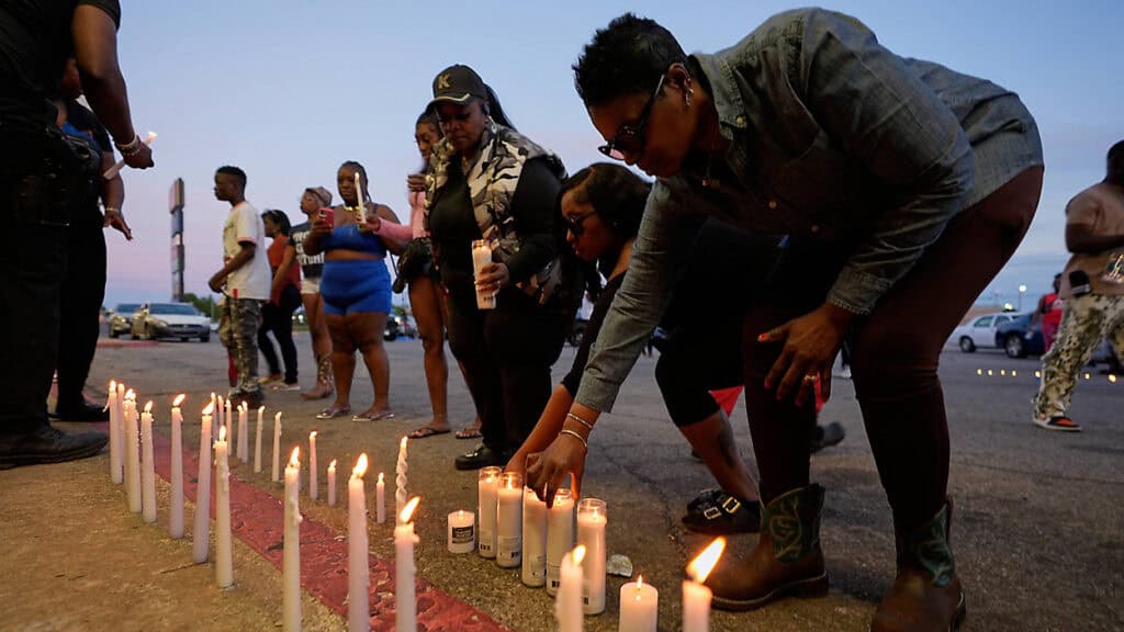 A woman places candles in a row on pavement as people gather at an evening vigil.