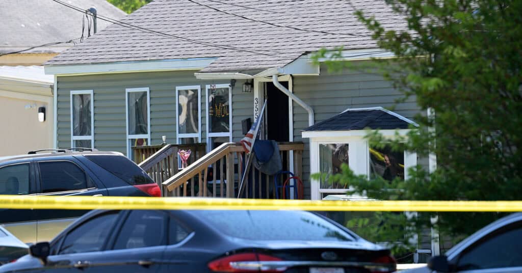 Yellow tape stretched in front of a two-story house with cars parked outside.