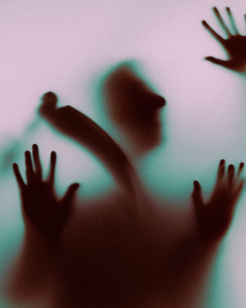 Silhouette of a person with hands pressed against a frosted glass panel, seen in pink and green light.