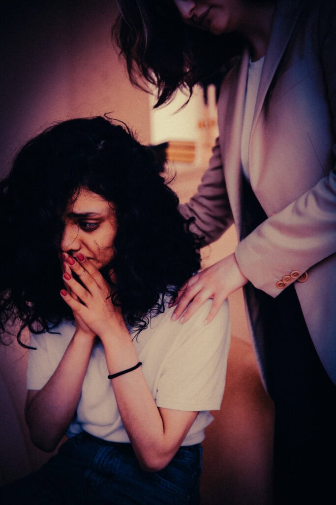 A woman with curly dark hair covers her mouth with her hands while another person rests a hand on her shoulder.