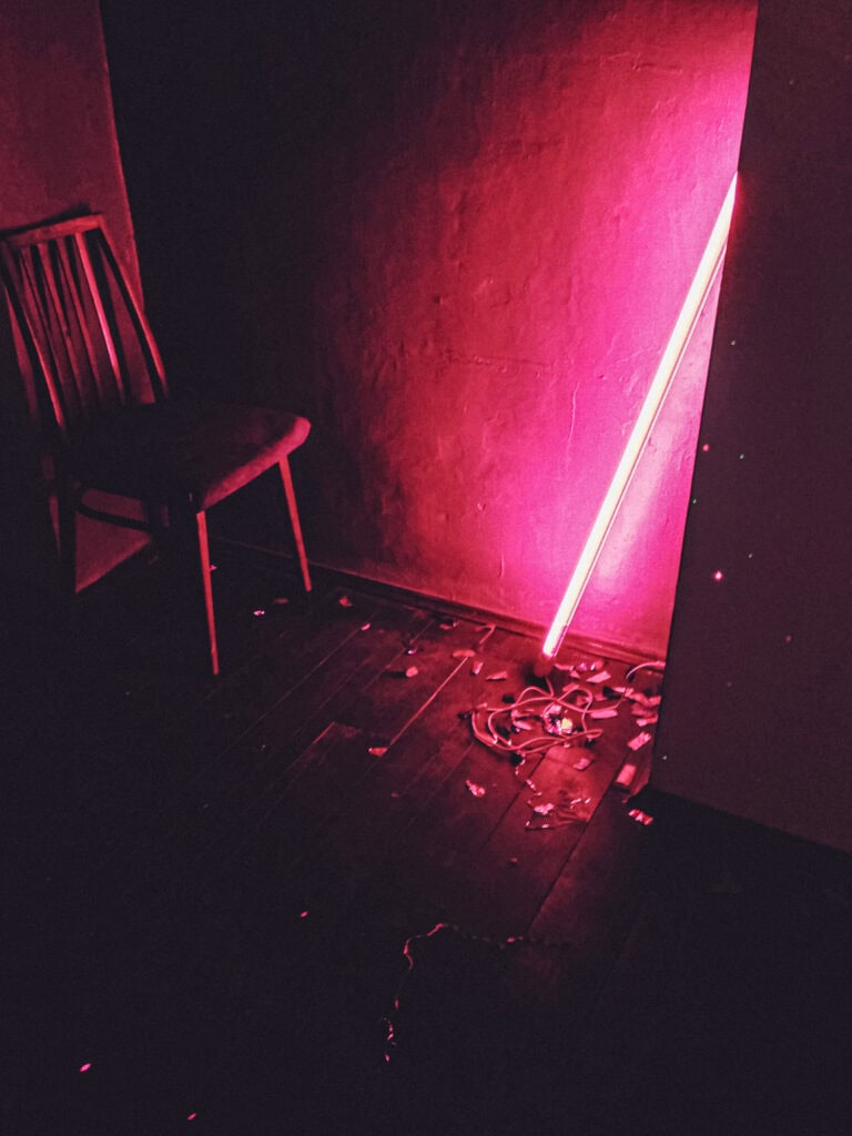 An empty wooden chair beside a wall lit by a leaning pink neon tube light.
