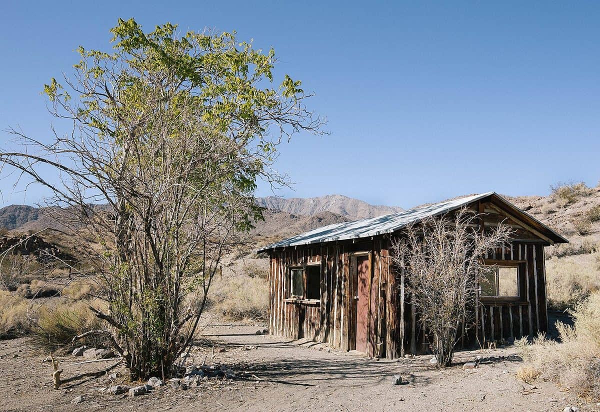 A weathered stone ranch building with a corrugated roof in a desert landscape under a blue sky