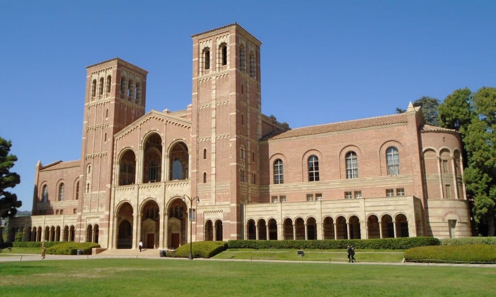 Royce Hall at UCLA, a red-brick Romanesque building with two tall arched bell towers and a long arcade, behind a green lawn under a clear blue sky.