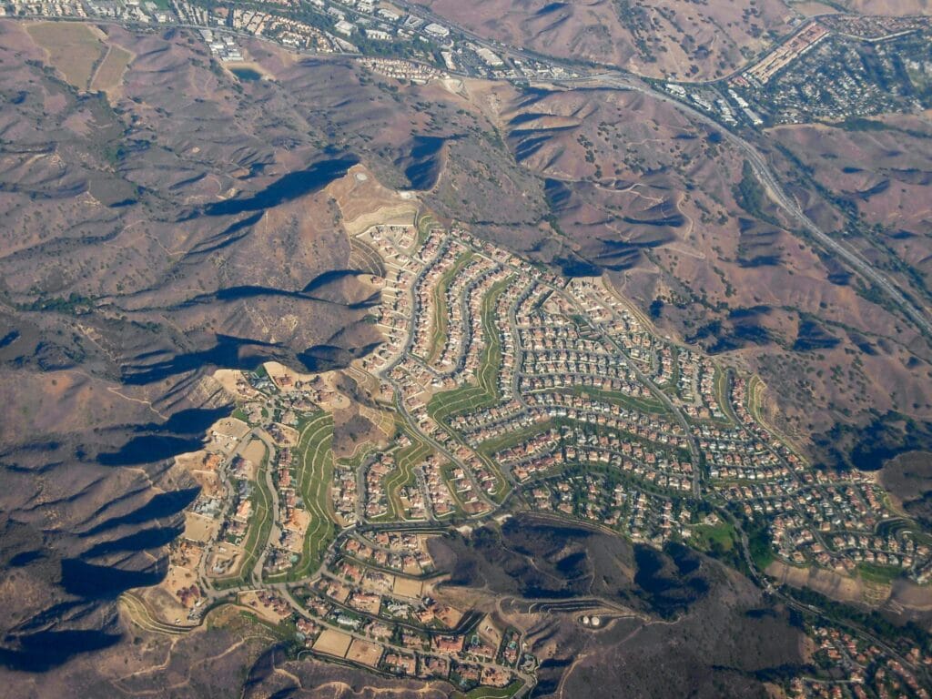 Aerial view of a hillside housing development in Calabasas, with curving streets of houses set among brown, sunlit mountain ridges and a freeway running along the top of the frame.