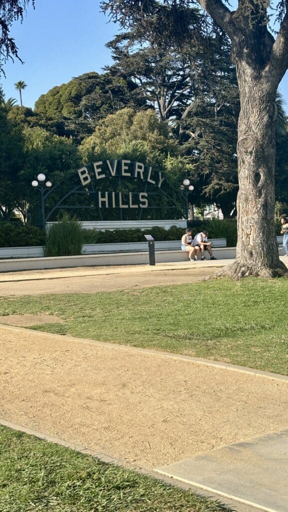 The white script Beverly Hills sign set in a lawn in a city park, with large shade trees and a gravel path in the foreground.