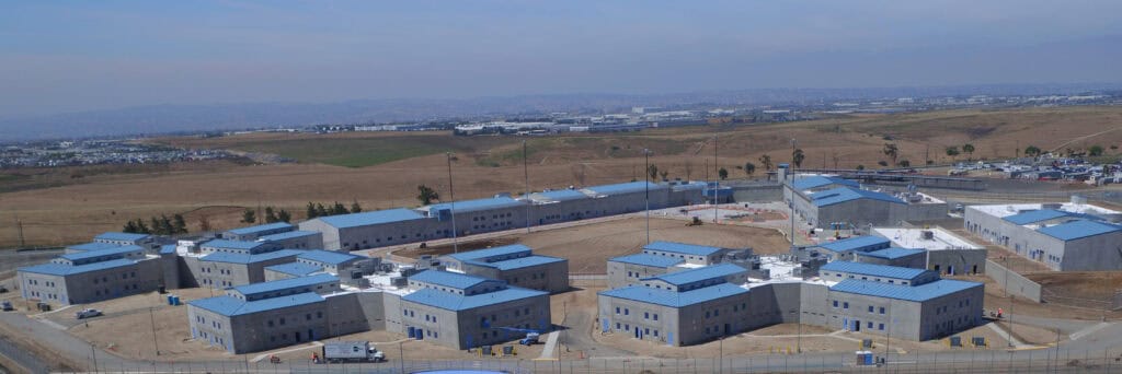 Elevated panoramic view of rows of low, flat-roofed prison buildings with pale blue roofs at Richard J. Donovan Correctional Facility, on flat, dry land.