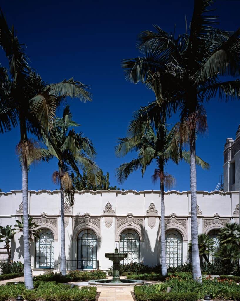 The white arcaded facade of Beverly Hills City Hall, framed by tall palm trees and a fountain under a deep blue sky.