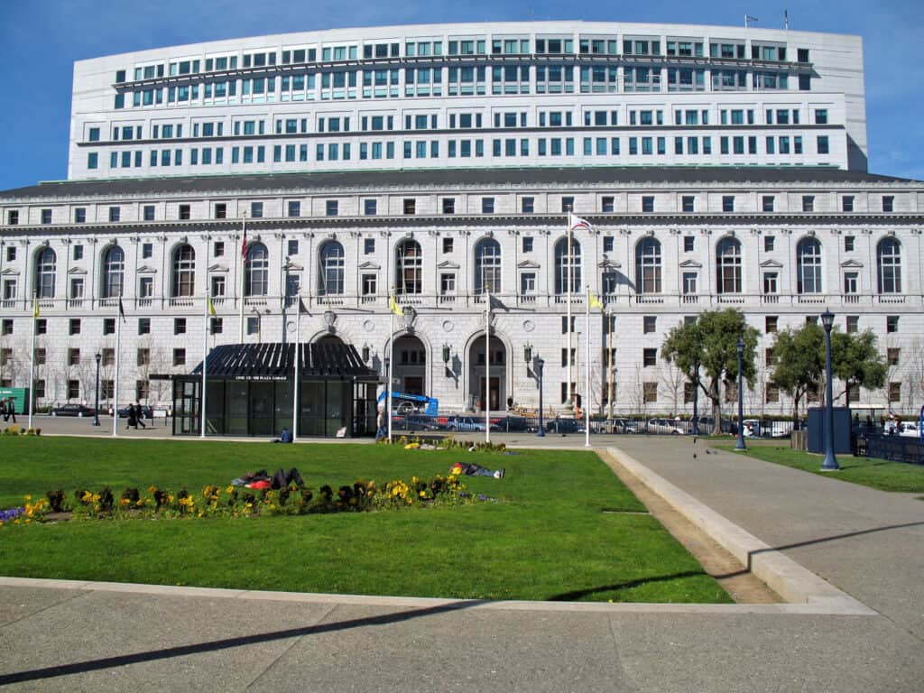 The pale granite Earl Warren Building in San Francisco's Civic Center, with a lawn and flower beds in front and a modern office tower behind.