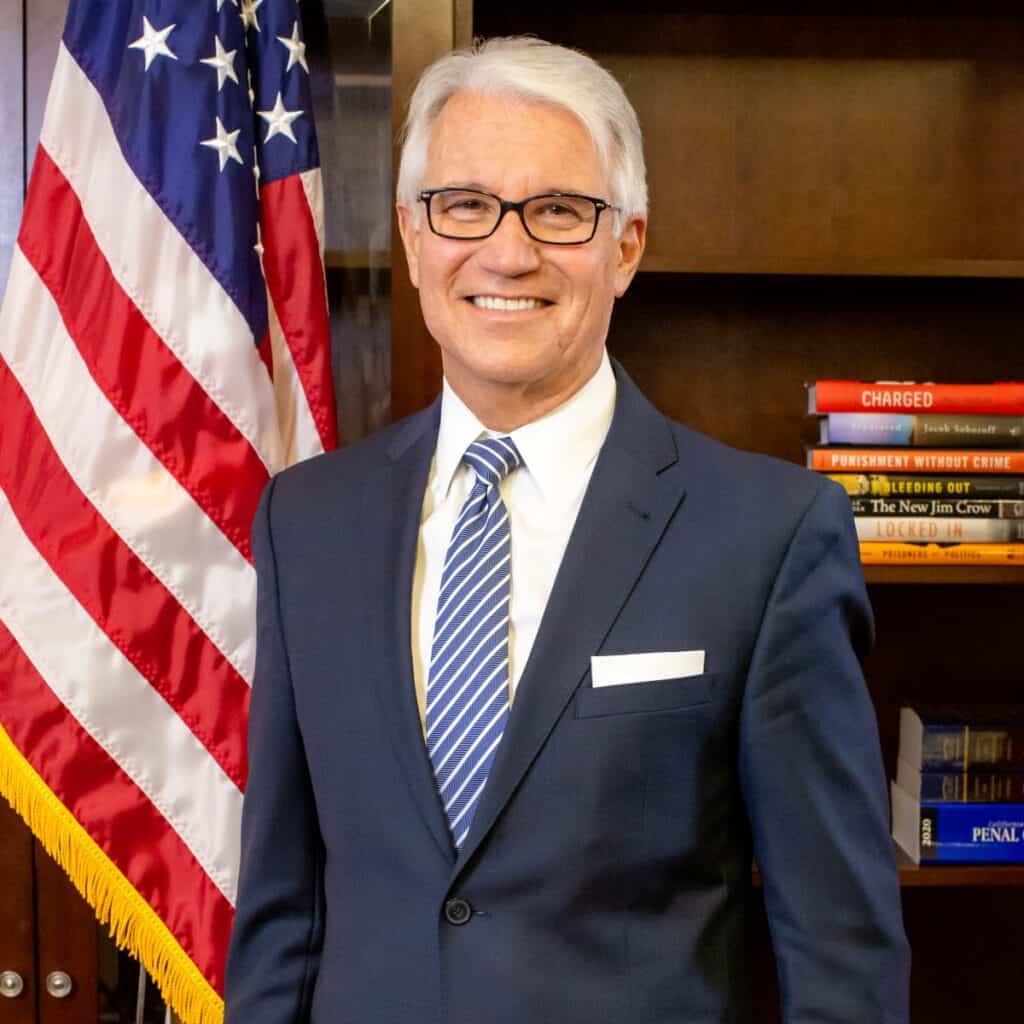 Official portrait of George Gascón, smiling, in a navy suit and striped tie, standing beside an American flag in front of a bookshelf.