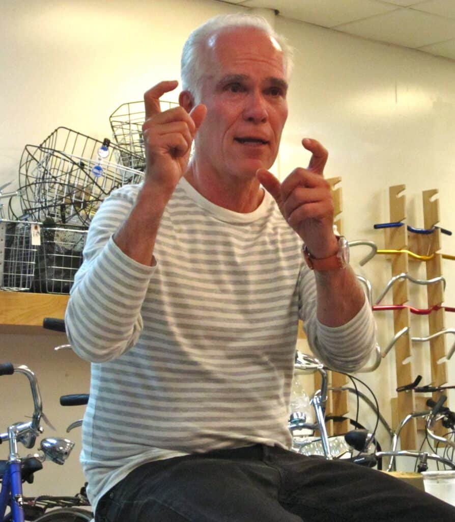 Gil Garcetti, a white-haired man in a striped sweater, gestures with both hands raised while speaking indoors, with bicycle wheels and racks behind him.