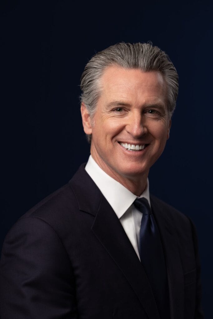 Official portrait of California Gov. Gavin Newsom, smiling, in a dark suit and tie against a dark blue background.