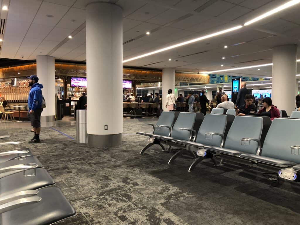 A bright, modern departure lounge at Los Angeles International Airport, with rows of gray seats, white columns, a polished floor and travelers walking past shops.