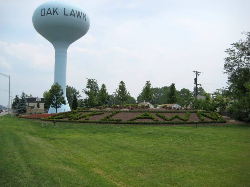 A white water tower painted OAK LAWN in blue letters stands beside a landscaped lawn and flower beds with trees, in a Chicago suburb.