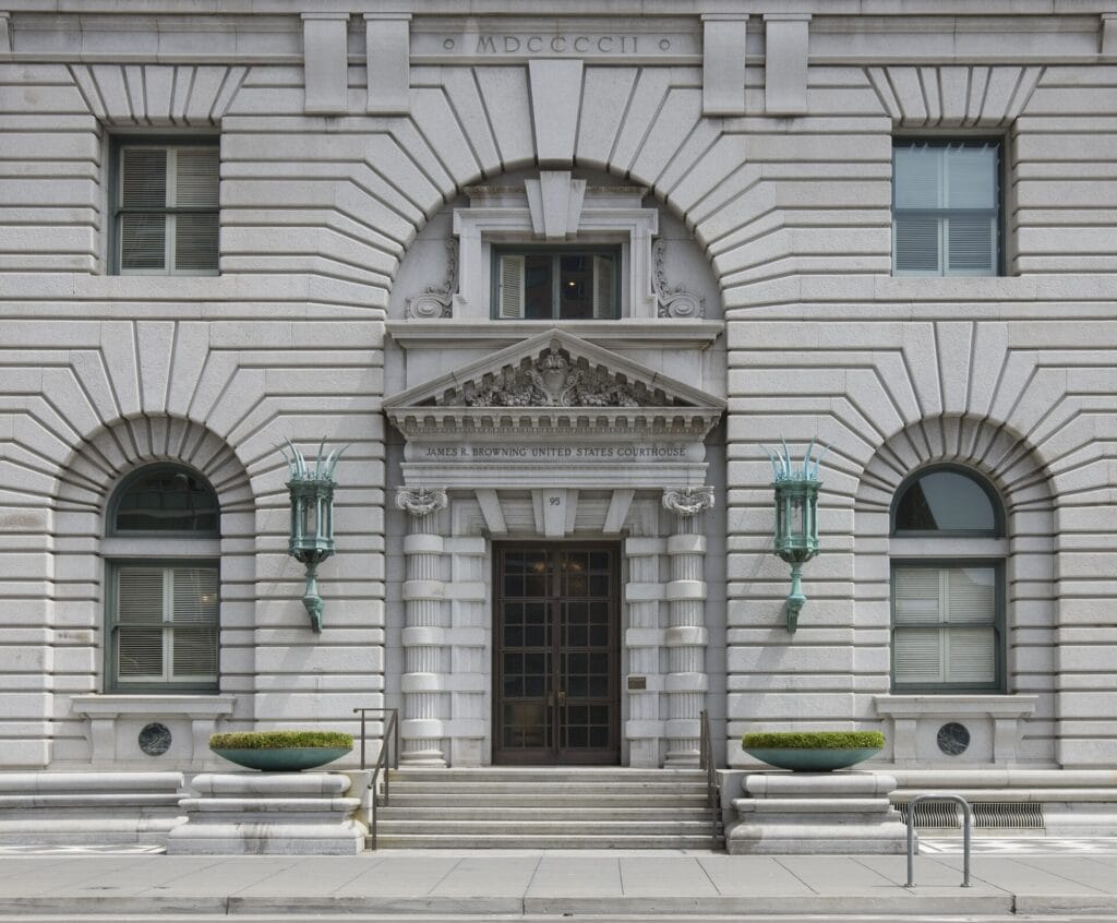 The ornate granite entrance of the James R. Browning U.S. Court of Appeals Building in San Francisco, with arched windows, carved stone detail and steps leading up to bronze doors.