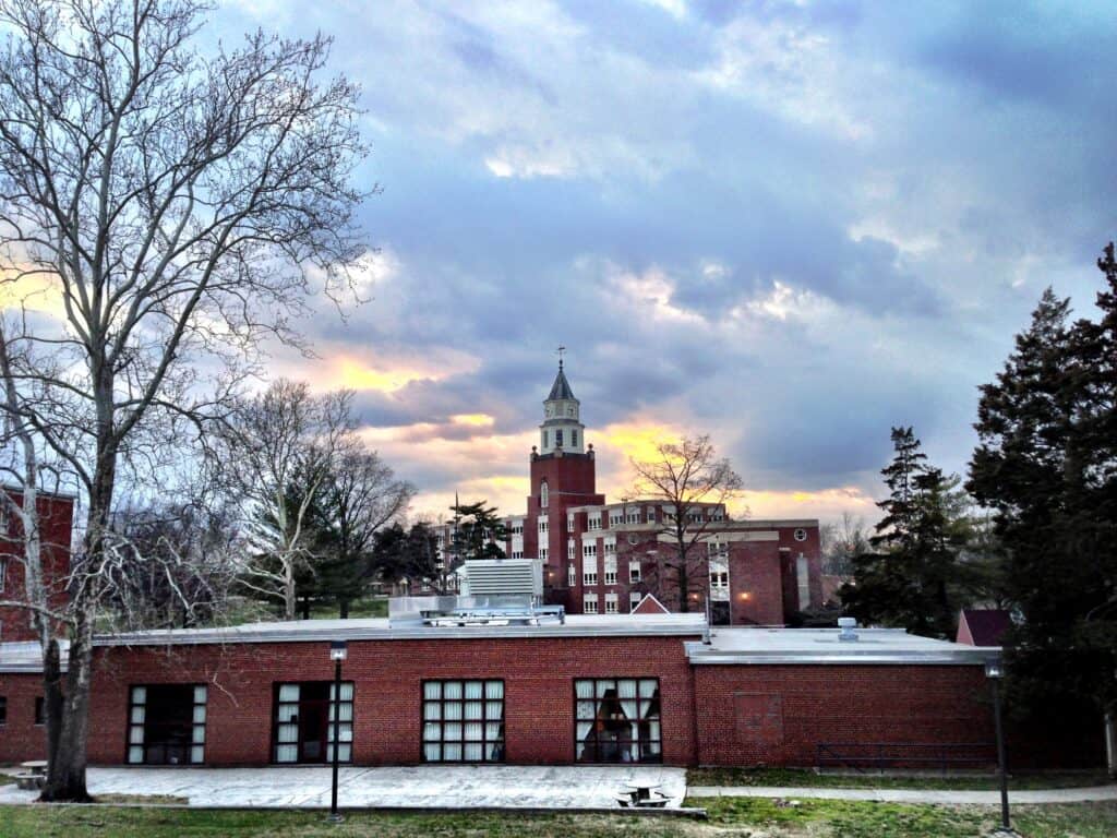 A red-brick university building with a white clock tower rising behind a long, low brick wing, under a cloudy sunset sky with bare trees in the foreground.